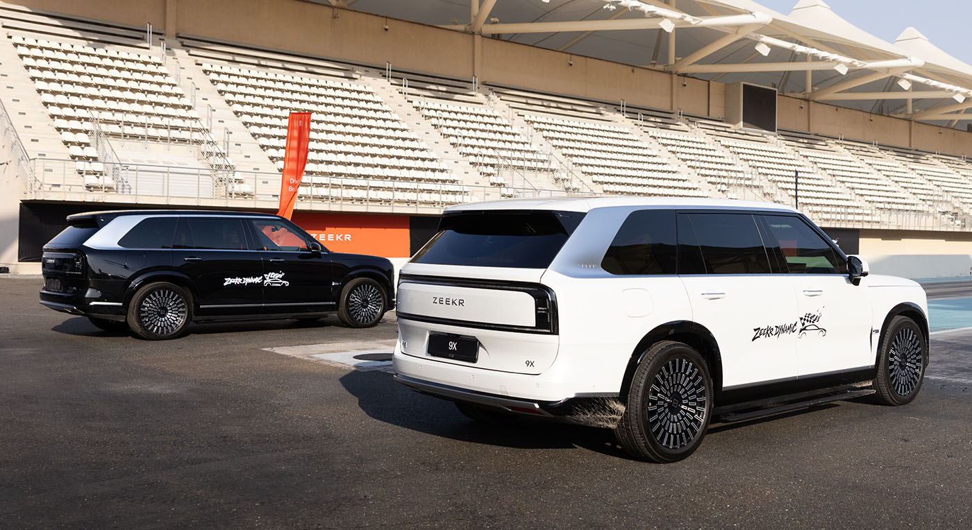 One black and one white luxury Zeekr 009 SUV parked on a race track tarmac in front of empty grandstands.
